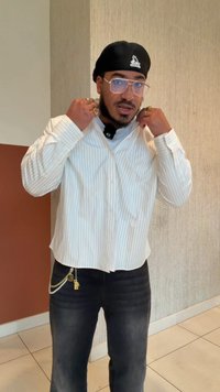 Camicia bianca a righe sottili con bottoni e maniche lunghe, abbinata a jeans scuri con una catena come accessorio. Indossati occhiali trasparenti e un berretto nero.