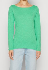 Maglietta verde a maniche lunghe realizzata in tessuto leggero, con ampio scollo a barca e una finitura leggermente testurizzata. Abbinata a jeans azzurri chiari.