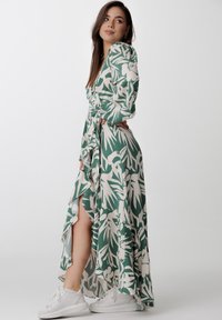 Zielona kwiatowa sukienka wrap z długimi rękawami, falbaniastym dołem i bocznym rozcięciem, w zestawieniu z białymi trampkami. Gładki materiał o lekkiej teksturze.