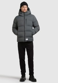 Grijze gewatteerde jas met een capuchon, voorzien van een voorzak en een rits aan de voorkant. Draag met zwarte broek en zwarte laarzen. Glad textuur.