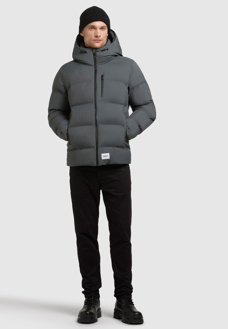 Grijze gewatteerde jas met een capuchon, voorzien van een voorzak en een rits aan de voorkant. Draag met zwarte broek en zwarte laarzen. Glad textuur.