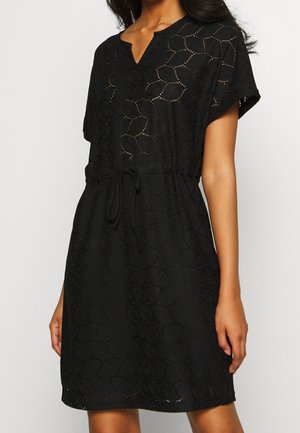 Vrouw die een zwarte kanten jurk draagt die tot aan de knie reikt, met korte mouwen, een V-hals en een taille met aantrekkoord.