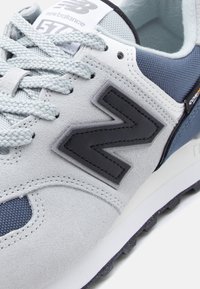 Pantof sport New Balance 574 gri și bleumarin, cu șireturi groase, din piele întoarsă și țesătură mesh, cu un logo negru „N” pe lateral.