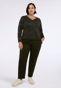 Kobieta z krótkimi ciemnymi włosami, ubrana w sweter w ciemnozielony wzór, pasujące spodnie i białe trampki, stojąca na jednolitym tle.