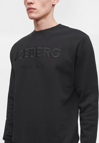 Jeune homme portant un sweat-shirt noir avec "ICEBERG since 1974" brodé sur le devant, coupé au niveau du cou et de la taille.
