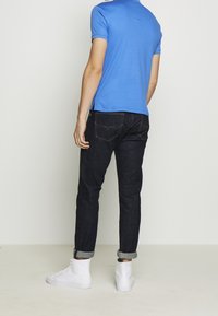 Mies, joka pukeutuu siniseen polo-paitaan, tummiin käärittyihin farkkuihin ja valkoisiin korkean varren tennareihin, seisoo yksinkertaista valkoista taustaa vasten.