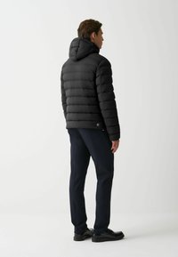 Črna puhovka s kapuco, z qiltano zasnovo in elastičnimi manšetami. Nosi se preko mornarsko modrih hlač in čevljev, gledano zadaj.