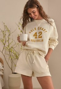 Ecrufarbenes Sweatshirt mit Waffelstruktur, versehen mit goldenen Buchstaben "GOLD COAST 42 AUSTRALIA." Kombiniert mit passenden Shorts, beide mit Kordelzug.