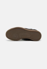 Zapatilla negra con una suela de goma texturizada, que presenta un patrón de huella marrón y el logo de Reebok. La parte superior muestra acentos de tela suave.