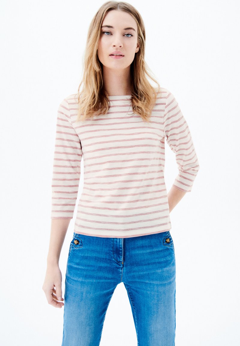 Gestreiftes Langarmshirt in hellem Rosa und Creme mit rundem Ausschnitt; kombiniert mit enganliegenden blauen Jeans, die dekorative Hardware aufweisen.