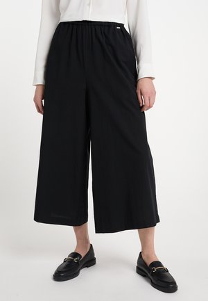 Musta leveälahkeinen housu, joissa on joustava vyötärö, valmistettu kevyestä kankaasta. Yhdistetty mustien nahkaisten loafer-kenkien kanssa. Tasainen rakenne.