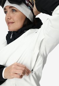 Veste de ski blanche avec des accents noirs, une ventilation zippée sous le bras, un col texturé et une surface lisse ; partiellement dézippée pour ajustement.