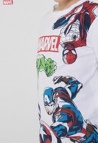 Biała koszulka z nadrukiem postaci Marvela: Spiderman, Hulk i Kapitan Ameryka w kolorowych wzorach. Okrągły dekolt, krótkie rękawy, luźny fason.