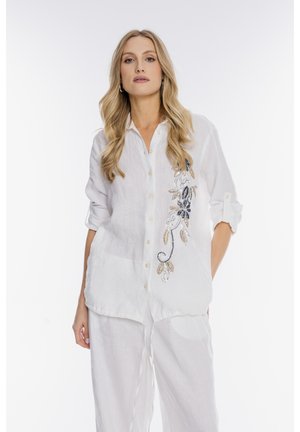 Blondynka w białej koszuli na guziki z kwiatowym haftem i dopasowanych luźnych białych spodniach, stojąca z jedną ręką w kieszeni.