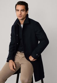 Uomo seduto su uno sgabello, indossa un cappotto navy, un maglione nero, una camicia grigia e pantaloni beige, contro uno sfondo grigio uniforme.