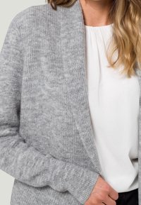 Cardigan grigio lavorato a maglia con apertura frontale, polsini a coste e texture morbida, indossato sopra una camicetta bianca, caratterizzato da un design rilassato.