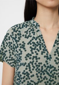 Blouse à manches courtes en tissu vert clair avec un col en V, présentant des motifs abstraits de points verts foncés et une coupe décontractée.