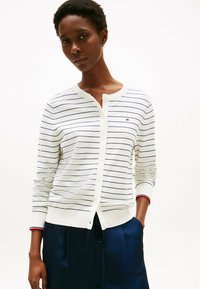 Cardigan bianco con strisce blu, chiusura con bottoni e polsini a coste con un accento rosso. Il tessuto appare leggero e liscio.