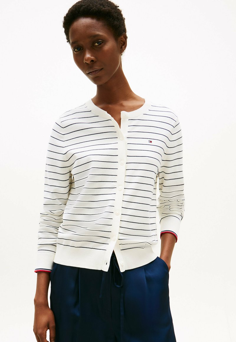 Cardigan bianco con strisce blu, chiusura con bottoni e polsini a coste con un accento rosso. Il tessuto appare leggero e liscio.