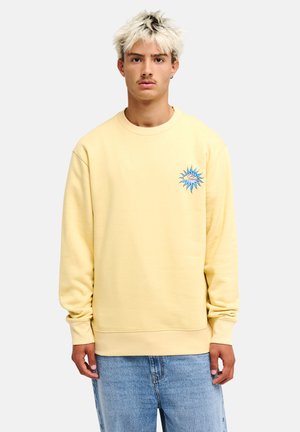 Jeune homme aux cheveux blonds portant un sweat-shirt jaune pâle avec un motif de soleil bleu sur la poitrine et un jean bleu sur un fond uni.
