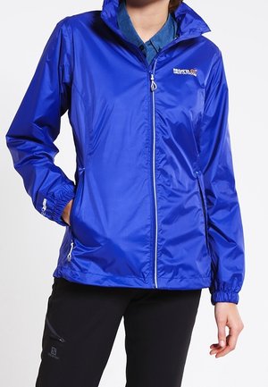 Person trägt eine hellblaue, leichte Jacke mit Reißverschluss, Kapuze, Taschen und Logos auf Brust und Ärmel, kombiniert mit schwarzen Hosen.