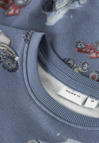 Zilā auduma džemperis ar traktoru un transportlīdzekļu apdrukām. Ribiņām apstrādāta apaļa kakla izgriezne, mīksta tekstūra un etiķete ar zīmolu "name it" un izmēru 92.