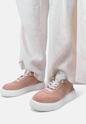 Lichtroze suède sneakers met dikke witte zolen en veters, gedragen bij losse witte broek, tegen een effen achtergrond.