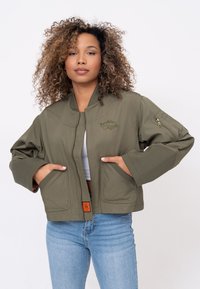 Olivgrüne Bomberjacke mit Reißverschluss, großen Taschen und einem Aufnäher auf der Brust. Verfügt über eine lockere Passform und Bündchen an den Ärmeln.