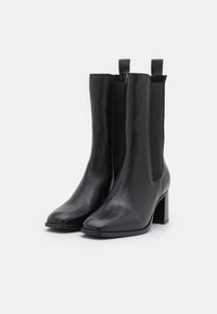 Bottes noires en cuir à cheville avec panneaux élastiques latéraux, bouts carrés et talons carrés. Texture lisse avec languettes pour faciliter l'enfilage.