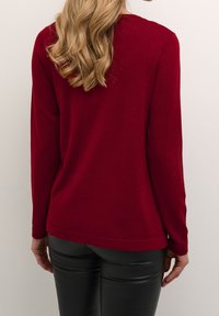 Bordeauxrode trui met lange mouwen, een gladde textuur en lichte ribbels bij de halslijn en zoom. Gecombineerd met zwarte leren broek.