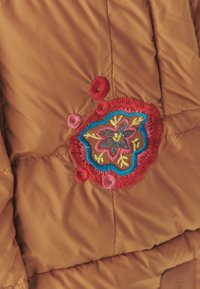 Brūns pūkains mētelis ar krāsainiem ziedu izšuvumiem, kuros ir sarkanas, rozā, zilas un zaļas akcenti, izceļot dažādas tekstūras un diegu izšuvumu detaļas.