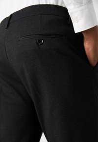 Gros plan sur une personne portant un pantalon de costume noir avec une poche arrière boutonnée et une chemise blanche avec le bouton de manchette visible.