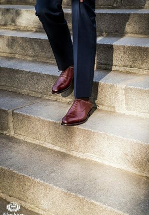 Braune Lederderby-Schuhe mit glatter Oberfläche und geraden Schnürsenkeln, getragen beim Herabsteigen von Steinstufen, kombiniert mit dunklen Hosen.