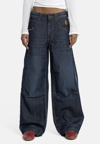 Oseba nosi temno modre široke kavbojke z gumbi in zadrgo, kombinirane z rdečimi čevlji in belim dolgotrajni zgornji del majice.