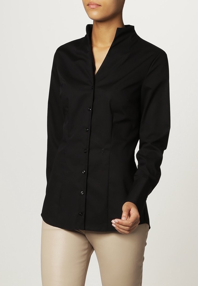 Femme portant un chemisier noir cintré boutonné avec un col montant et un pantalon beige, posant avec une main légèrement levée.