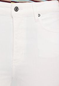 Gros plan sur un jean en denim blanc montrant le bouton avant, la boucle de ceinture et la poche avec rivet métallique sur un fond en tissu rayé.