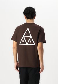 Camiseta de algodón marrón con mangas cortas. Logotipo triangular blanco con las letras "H", "U" y "F" en la parte posterior. Textura suave, ajuste estándar.