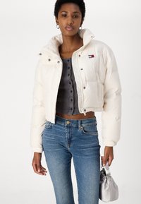 Giacca puffer cropped color crema con colletto alto, bottoni a pressione e un patch con logo. Abbinata a una camicia grigia con bottoni e jeans blu.