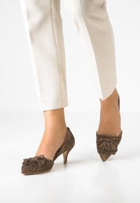 Jambes dans un pantalon beige portant des chaussures à talons kitten en suède marron avec des détails en franges sur un sol et un fond blancs.