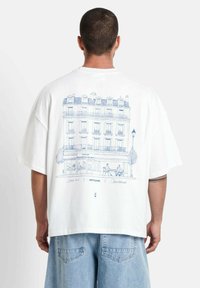 Witte katoenen t-shirt met een blauwe lijntekening van een Parijse gebouw met buitenzitplaatsen, korte mouwen en een ontspannen pasvorm.