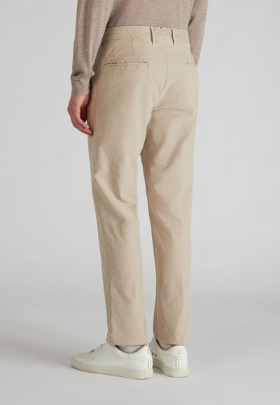 Pantalones beige de corte relajado y textura sutil, con bolsillos traseros y combinados con zapatillas blancas. Diseño liso, sin patrones visibles.