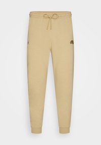 Beige Baumwoll-Jogginghosen mit elastischem Bund und Kordelzug, versehen mit einem kleinen schwarzen Logo und einem bedruckten Detail am rechten Oberschenkel.