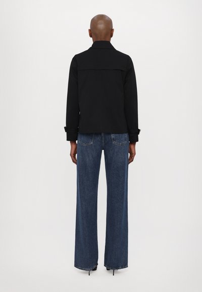 Veste noire avec col et poignets boutonnés, associée à un pantalon en denim bleu à jambes larges, avec une finition décolorée et des poches arrière visibles.