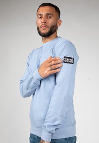 Sudadera azul claro hecha de una mezcla de algodón, con puños y dobladillo acanalados, y un parche negro etiquetado "WRONG FRIENDS" en la manga izquierda.