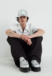 Strieborné topánky, čierne široké nohavice a biele polo s zipsovým golierom. Svetlosivá čiapka dopĺňa vzhľad. Model sedí na podlahe.