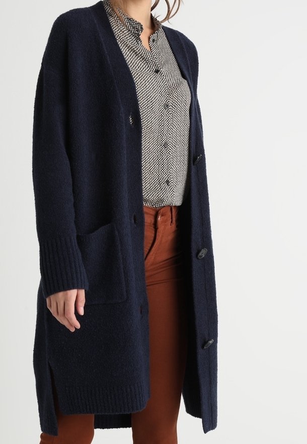 Tmavě modrý dlouhý kardigan z měkké látky, s žebrovanými manžetami, postranními kapsami a velkými knoflíky, vrstvený přes vzorovanou košili.