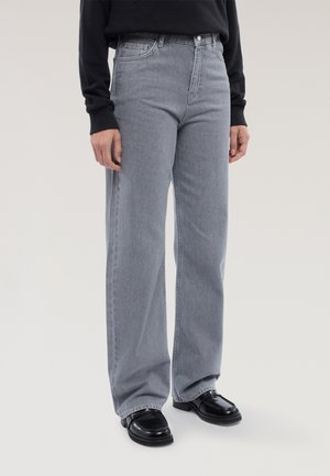 Persoon draagt grijze wijde jeans met hoge taille, zwart langarmshirt en zwarte leren loafers, staand tegen een effen achtergrond.