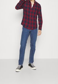 Rød og marineblå ternet flannel skjorte med lange ærmer, parret med blå jeans og sorte sneakers. Håret stylet bagud, arme hævet.