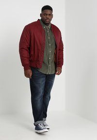 Giacca bomber bordeaux, camicia verde oliva con bottoni, jeans blu scuro e sneaker navy. Texture morbide con una vestibilità rilassata.