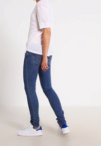 Top blanco de manga corta con puños enrollados, combinado con jeans ajustados azul oscuro y zapatillas blancas con acentos azules, que muestran una silueta elegante.
